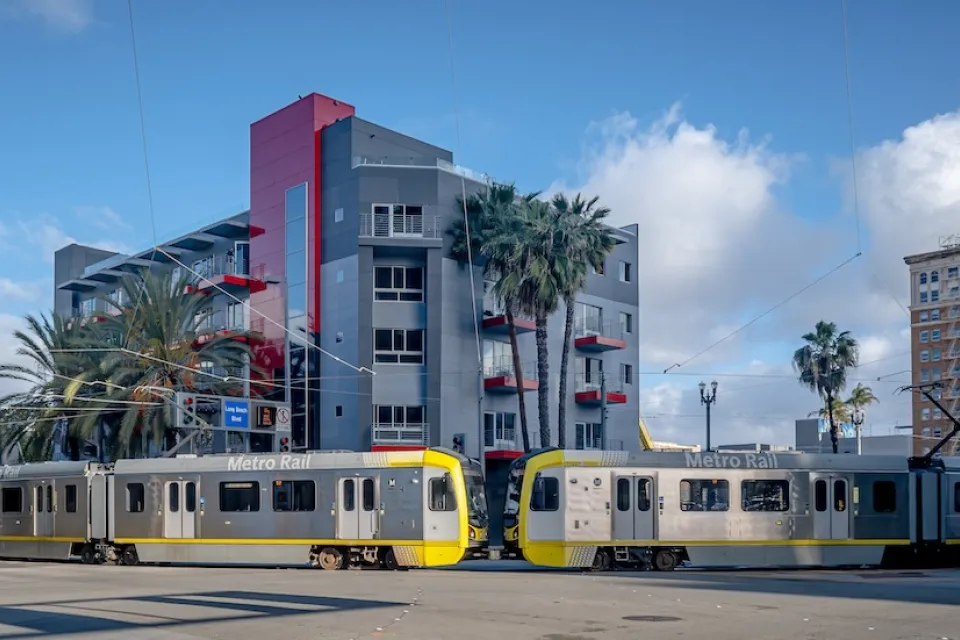 Two silver trains leaving station in Long Beach, California in front of mid-rise residential building.